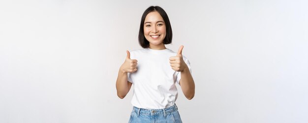 Feliz hermosa chica coreana mostrando los pulgares hacia arriba en señal de aprobación sonriendo complacida como algo recomendando pararse sobre fondo blanco