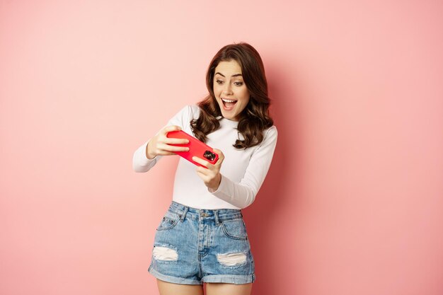 Feliz hermosa chica jugando videojuegos móviles, sosteniendo el teléfono inteligente horizontalmente, mirando en el celular con cara emocionada, fondo rosa.