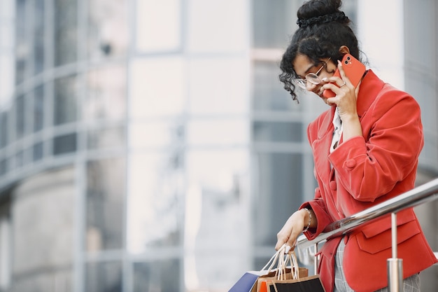 Feliz hermosa joven con gafas, con bolsas de la compra, hablando por un teléfono móvil de un centro comercial