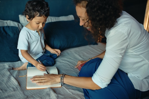 Feliz hermosa joven latina sentada en la cama con su pequeño y lindo hijo, colocando la mano sobre el papel y trazando sus contornos con lápiz.