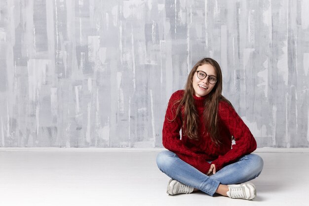 Feliz hermosa joven mujer caucásica con anteojos, jeans, suéter acogedor y zapatillas de deporte sentado con las piernas cruzadas en el piso y mirando con amplia sonrisa alegre, shwing dientes blancos