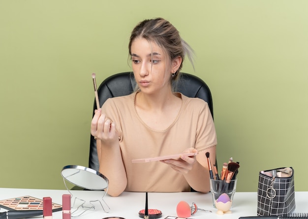 Feliz hermosa joven sentada en el escritorio con herramientas de maquillaje aplicando sombra de ojos con pincel de maquillaje aislado en la pared verde oliva