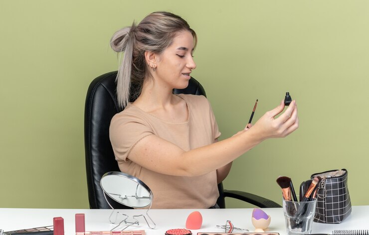 Foto gratuito feliz hermosa joven sentada a la mesa con herramientas de maquillaje sosteniendo rímel en la cámara aislada en la pared verde oliva