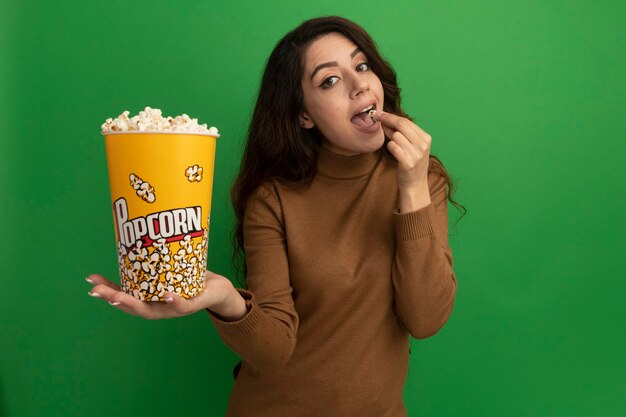 Feliz hermosa joven sosteniendo y probando un cubo de palomitas de maíz aislado en la pared verde