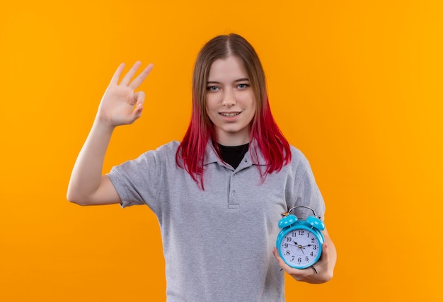 Feliz hermosa joven vestida con camiseta gris con reloj despertador mostrando gesto bien sobre fondo amarillo aislado