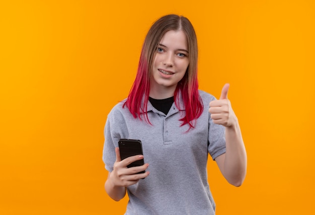 Feliz hermosa joven vestida con camiseta gris sosteniendo el teléfono con el pulgar hacia arriba sobre fondo amarillo aislado