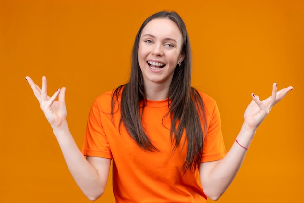 Feliz hermosa joven vestida con camiseta naranja sonriendo alegremente positiva y feliz de pie con los brazos levantados sobre fondo naranja aislado