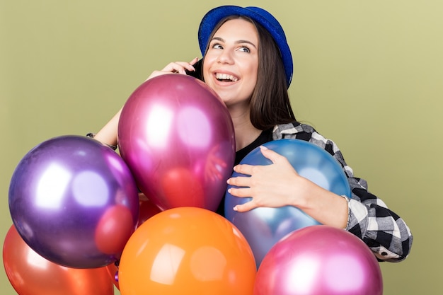 Feliz hermosa joven vestida con sombrero azul de pie detrás de globos habla por teléfono