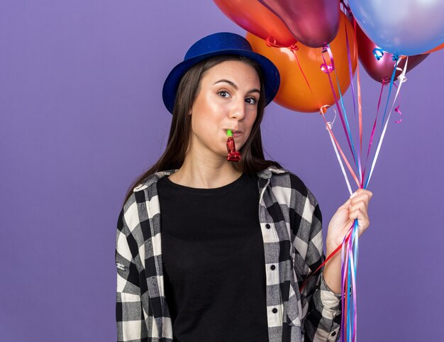 Feliz hermosa joven vistiendo gorro de fiesta sosteniendo globos soplando silbato de fiesta