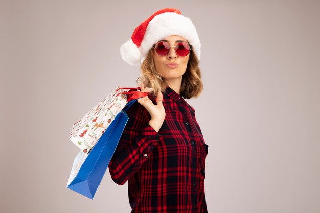 Feliz hermosa joven vistiendo gorro de navidad con gafas sosteniendo una bolsa de regalo en el hombro aislado sobre fondo blanco.