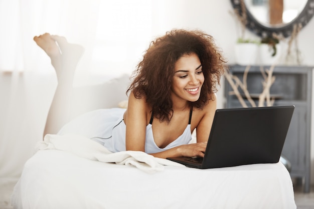 Feliz hermosa mujer africana en ropa de dormir acostado en cama en casa sonriendo mirando portátil.