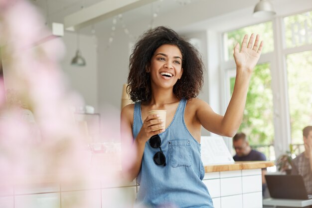 Feliz hermosa mujer africana sentada en la cafetería sonriendo riendo saludo bebiendo café.