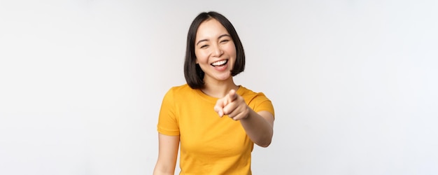 Feliz hermosa mujer asiática riendo señalando con el dedo a la cámara y riéndose sonriendo despreocupada de pie en camiseta amarilla sobre fondo blanco.