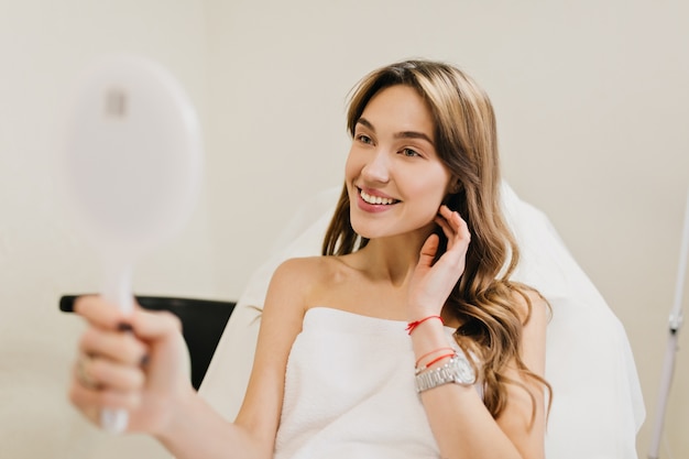 Feliz hermosa mujer con cabello largo morena después de la terapia de cosmetología sonriendo al espejo en la habitación blanca. Alegría, felicidad, buenos resultados, verdaderas emociones positivas