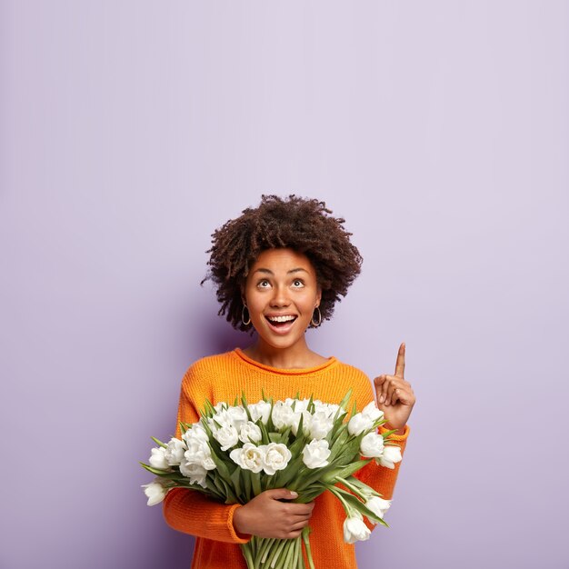 Feliz hermosa mujer con cabello nítido, puntos arriba en el espacio de la copia, sonríe tiernamente, sostiene tulipanes blancos, está de buen humor