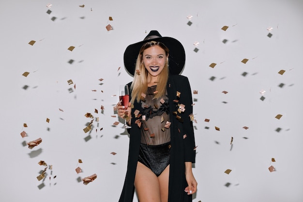 Feliz hermosa mujer con cabello rubio posando con vino sobre pared aislada con confeti