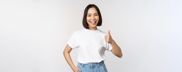 Feliz hermosa mujer coreana sonriendo complacida mostrando los pulgares hacia arriba en aprobación recomendando marca o empresa de pie sobre fondo blanco.
