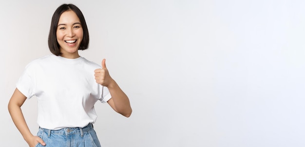 Feliz hermosa mujer coreana sonriendo complacida mostrando Thumbs up en aprobación recomendando marca o com