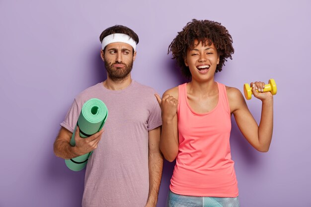 Feliz hermosa mujer con corte de pelo afro apunta con el pulgar al marido con expresión insatisfecha, tienen un entrenamiento activo juntos, usan ropa deportiva. Chico molesto no está ansioso por tener entrenamiento físico