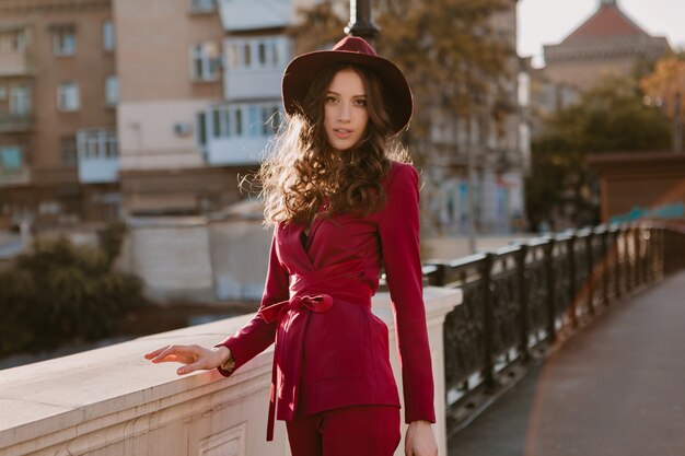 Feliz hermosa mujer elegante en traje púrpura caminando en las calles de la ciudad, tendencia de moda primavera verano otoño temporada con sombrero, sosteniendo el bolso