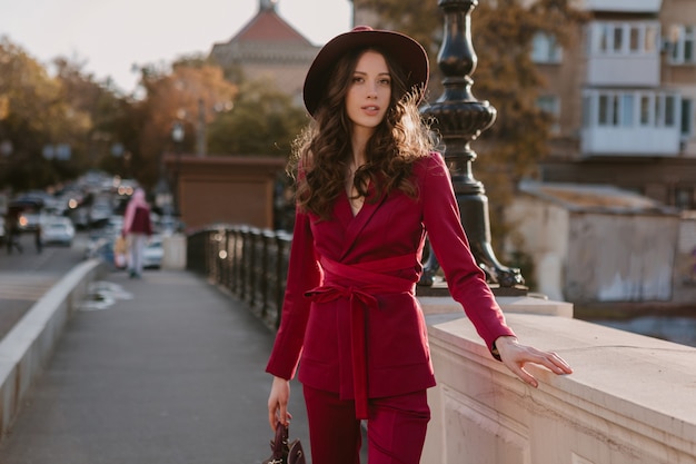 Feliz hermosa mujer elegante en traje púrpura caminando en las calles de la ciudad, tendencia de moda primavera verano otoño temporada con sombrero, sosteniendo el bolso