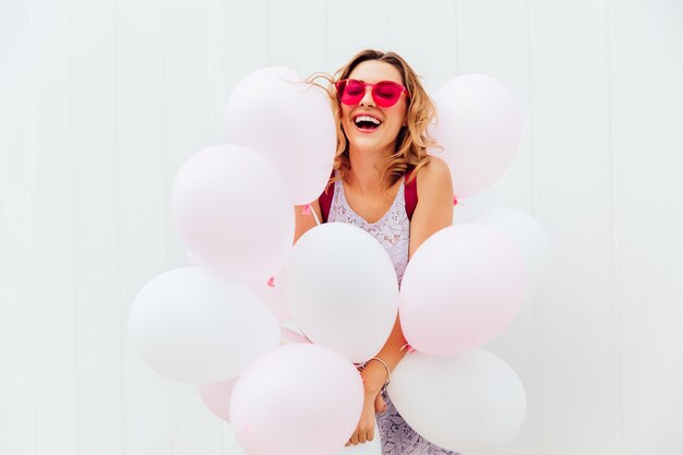 Feliz hermosa mujer joven en gafas de sol de color rosa tiene globos blancos, sonriendo alegremente