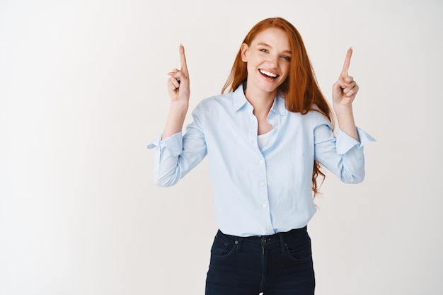 Feliz hermosa mujer pelirroja apuntando con el dedo hacia arriba y riendo, sonriendo al frente, vistiendo jeans y camisa de cuello azul