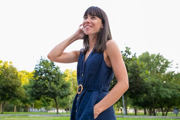 Feliz hermosa mujer de pelo negro de pie en el parque de la ciudad, mirando a otro lado y sonriendo. Señora disfrutando del tiempo libre al aire libre en verano. Plano medio, ángulo bajo. Concepto de retrato femenino