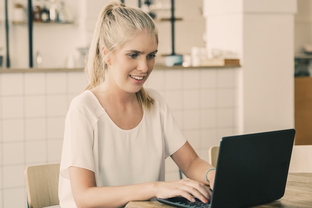 Feliz hermosa mujer rubia sentada a la mesa en el espacio de trabajo conjunto, usando la computadora portátil, mirando la pantalla y sonriendo