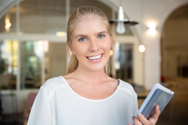 Feliz hermosa mujer rubia vestida con camisa blanca, de pie en el espacio de trabajo conjunto, sosteniendo el bloc de notas con papeles, posando