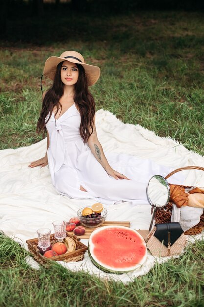 Feliz hermosa mujer con sombrero de ala y vestido blanco mientras está de pie y sosteniendo una canasta de pan en el parque de verano