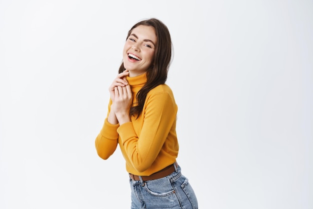 Feliz hermosa mujer en suéter amarillo riendo y expresando emociones positivas sonriendo a la cámara de pie medio vuelta sobre fondo blanco.