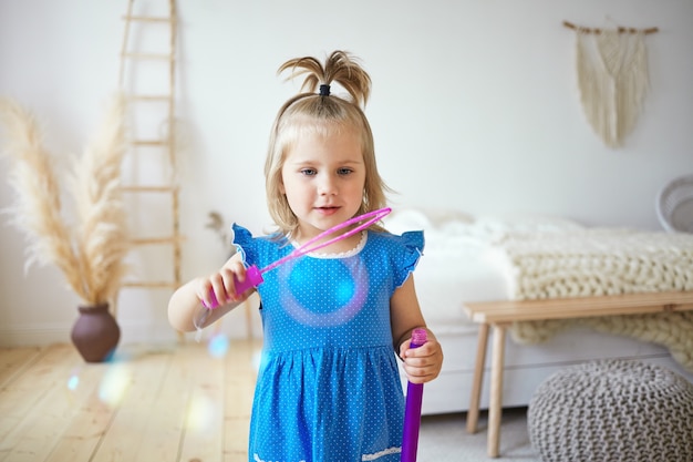 Feliz hermosa niña con cola de caballo divirtiéndose en el interior, soplando pompas de jabón en el dormitorio de los padres. Encantadora niña vistiendo un lindo vestido azul entreteniéndose, jugando solo en casa