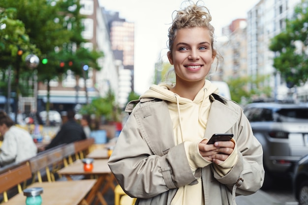 Feliz hermosa niña esperando a alguien fuera de la cafetería, sosteniendo el teléfono móvil y sonriendo.