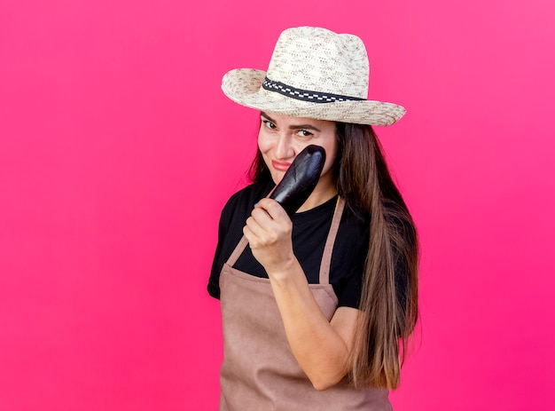 Feliz hermosa niña jardinero en uniforme con sombrero de jardinería sosteniendo berenjena aislado en rosa