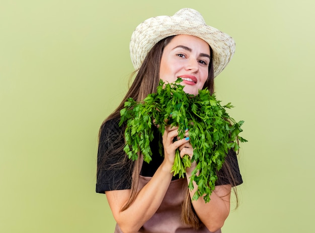 Feliz hermosa niña jardinero en uniforme con sombrero de jardinería sosteniendo cilantro alrededor de la cara aislada sobre fondo verde oliva