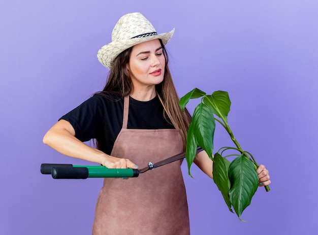 Feliz hermosa niña jardinero en uniforme con sombrero de jardinería sosteniendo y cortando plantas con tijeras aisladas en azul