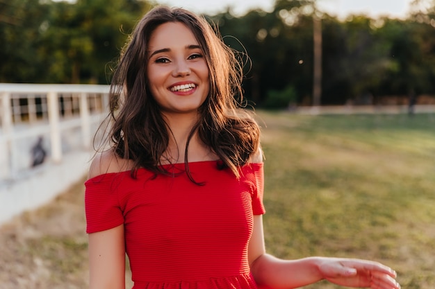 Feliz hermosa niña de pie en la naturaleza borrosa con una sonrisa. Mujer morena soñadora en vestido rojo posando con risa sincera