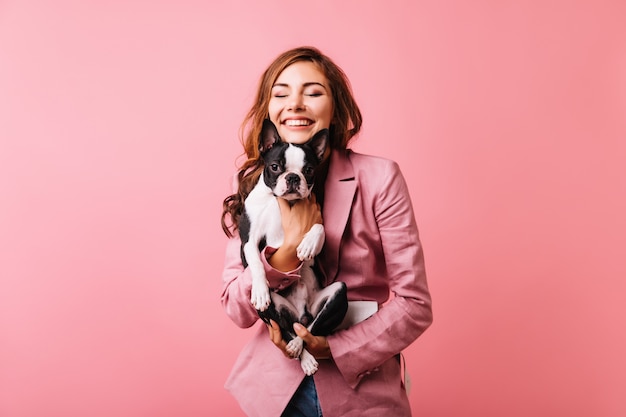 Feliz hermosa niña sosteniendo bulldog francés y sonriendo con los ojos cerrados. Retrato de mujer pelirroja romántica con cachorro aislado en rosa.