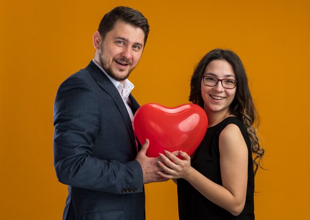 Feliz y hermosa pareja hombre y mujer con globo rojo en forma de corazón abrazando celebrando el día de San Valentín sobre pared naranja