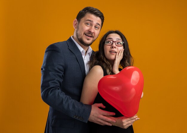 Feliz y hermosa pareja hombre y mujer con globo rojo en forma de corazón abrazando celebrando el día de San Valentín sobre pared naranja