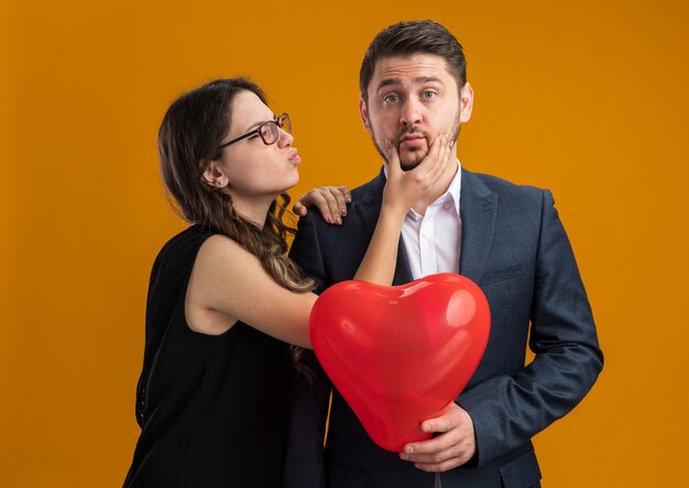 Feliz y hermosa pareja hombre y mujer con globo rojo en forma de corazón feliz enamorado celebrando el día de San Valentín sobre pared naranja