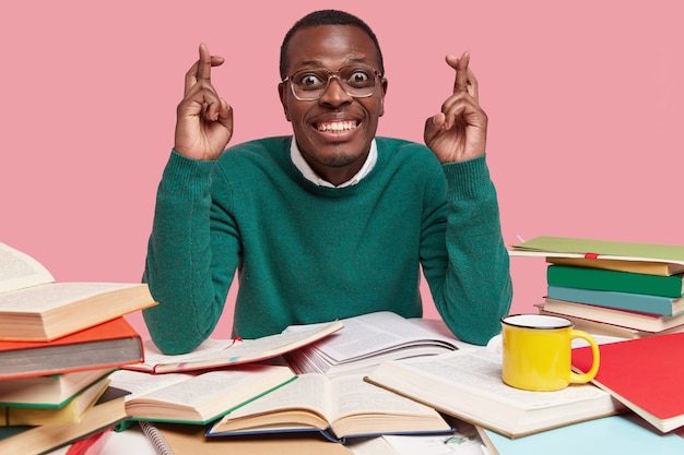 Foto gratuita feliz hombre afroamericano con amplia sonrisa, dientes blancos mantiene los dedos cruzados, cree en la buena suerte, lee literatura, bebe té caliente de una taza amarilla