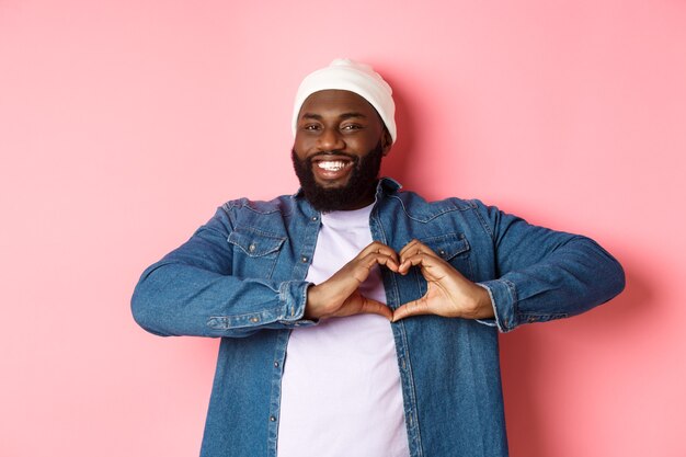 Feliz hombre afroamericano mostrando el signo del corazón, te amo gesto, sonriendo a la cámara, fondo rosa