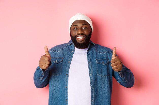 Feliz hombre afroamericano que muestra el pulgar hacia arriba en aprobación, como algo bueno, alabando mucho, de pie sobre fondo rosa