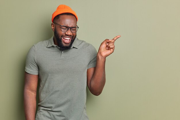 Feliz hombre afroamericano se ríe alegremente apunta lejos muestra algo fresco y divertido viste sombrero y camiseta casual gafas transparentes posa en el estudio demuestra espacio de copia