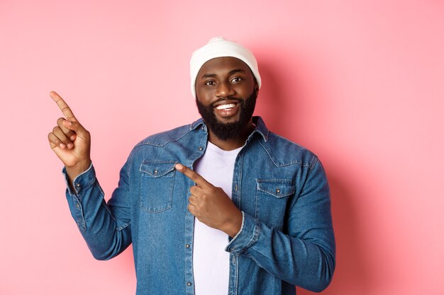 Feliz hombre afroamericano señalando con el dedo en la esquina superior izquierda, mostrando el logotipo de la oferta promocional, sonriendo complacido, vistiendo gorro con chaqueta vaquera, fondo rosa