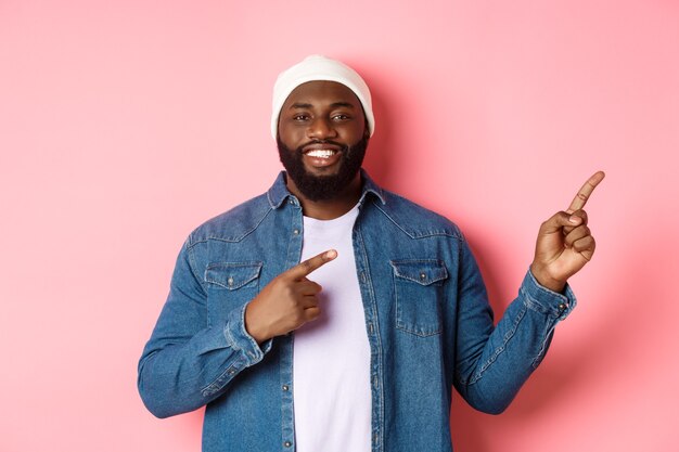 Feliz hombre afroamericano sonriendo, señalando con el dedo a la derecha y mostrando promo, haciendo un anuncio, de pie sobre un fondo rosa.