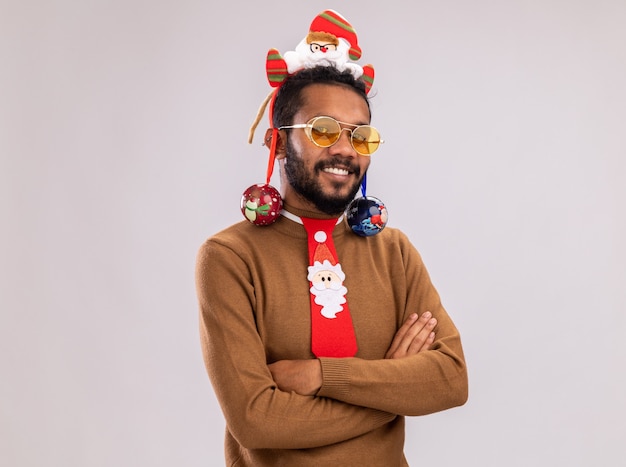 Foto gratuita feliz hombre afroamericano en suéter marrón y santa rim en la cabeza con divertida corbata roja sosteniendo bolas de navidad mirando a cámara con una sonrisa en la cara de pie sobre fondo blanco.