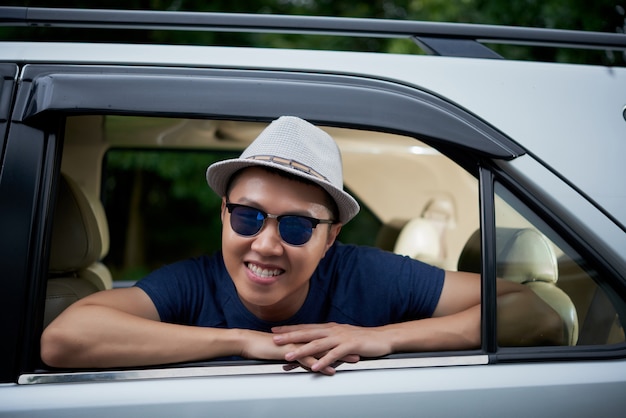 Feliz hombre asiático con sombrero y gafas de sol posando en la ventana trasera del coche